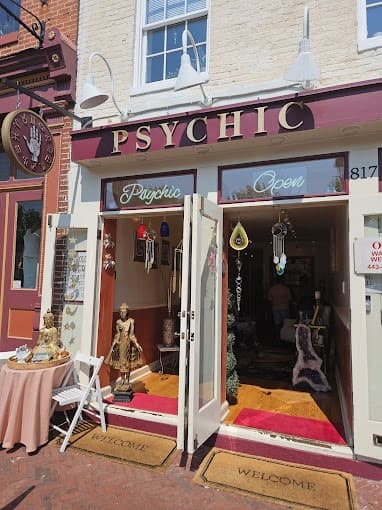 The storefront of a psychic and spiritual services business displays a maroon sign with gold letters spelling "PSYCHIC," an illuminated "Psychic Open" sign, and an open door revealing various spiritual and mystical items inside, with a small table holding decorative statues and a welcome mat outside.