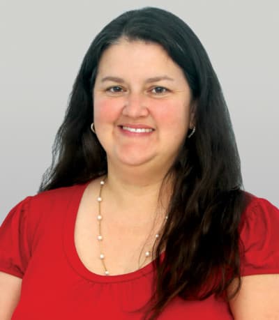 A woman with long dark hair, wearing a red top and a pearl necklace, smiling against a plain light background.
