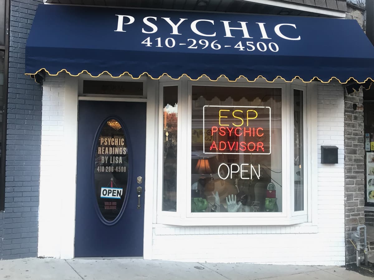 The storefront features a blue awning with the word "PSYCHIC" and a phone number, a door with a sign advertising psychic readings by Lisa, and a window displaying a neon sign that reads "ESP PSYCHIC ADVISOR" and "OPEN," along with various psychic-related items inside.